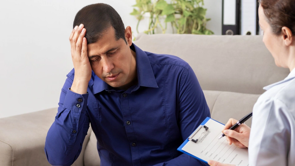 “A man sits with his head in his hand during a therapy session while a clinician takes notes on a clipboard, representing symptoms of PTSD
