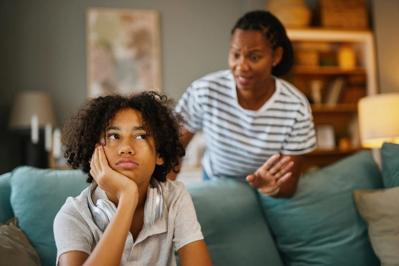 A teenager sits on a couch looking frustrated and disengaged while an adult speaks to them in a home setting