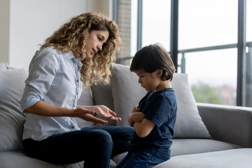 A mother speaks calmly to a young child who sits with arms crossed on a couch, appearing upset or resistant during a conversation at home