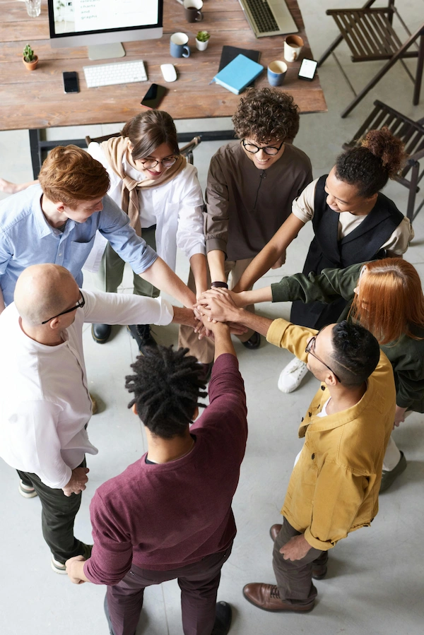 A diverse group of people stand in a circle with their hands stacked together, symbolizing unity, inclusion, and racial identity.