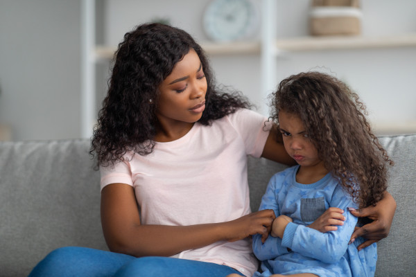 A mother sits beside her young child on a couch, offering comfort while the child sits with arms crossed and appears upset.