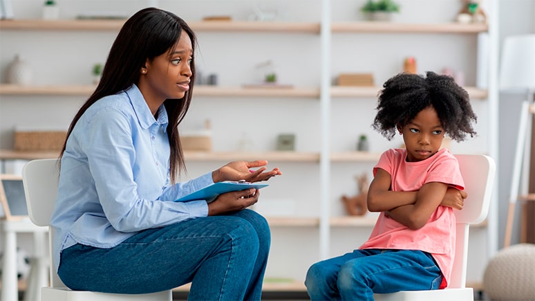 A therapist speaks gently to a young child who sits with arms crossed, appearing upset or resistant during a counseling session.